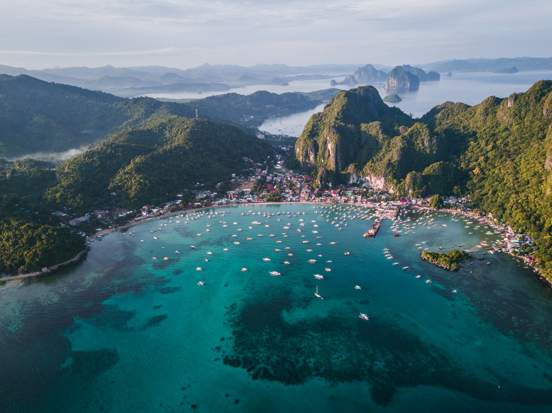 Vista aérea de uma enseada de águas turquesa cercada por morros verdes, com dezenas de barcos ancorados
